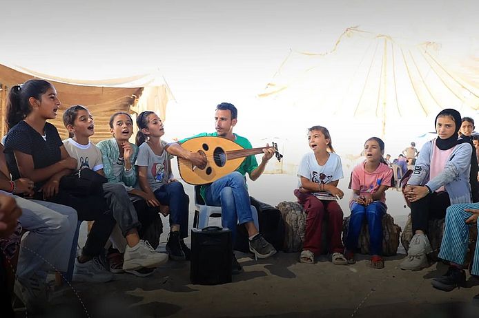Kinder singen im Kreis neben einem Mann mit einem Instrument