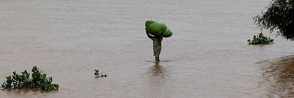 Ein Mann trägt einen großen Sack auf dem Kopf durch eine überflutete Landschaft. 