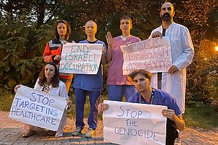 Menschen in Kittel und Notarzt-Jacke zeigen Schilder in Solidarität mit Gesundheitsarbeiter:innen in Gaza.