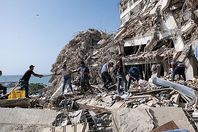 Mit einer Menschenkette tragen freiwillige Helfer:innen und Angehörige Schutt ab, um Menschen aus einem eingestürzten Haus zu befreien.